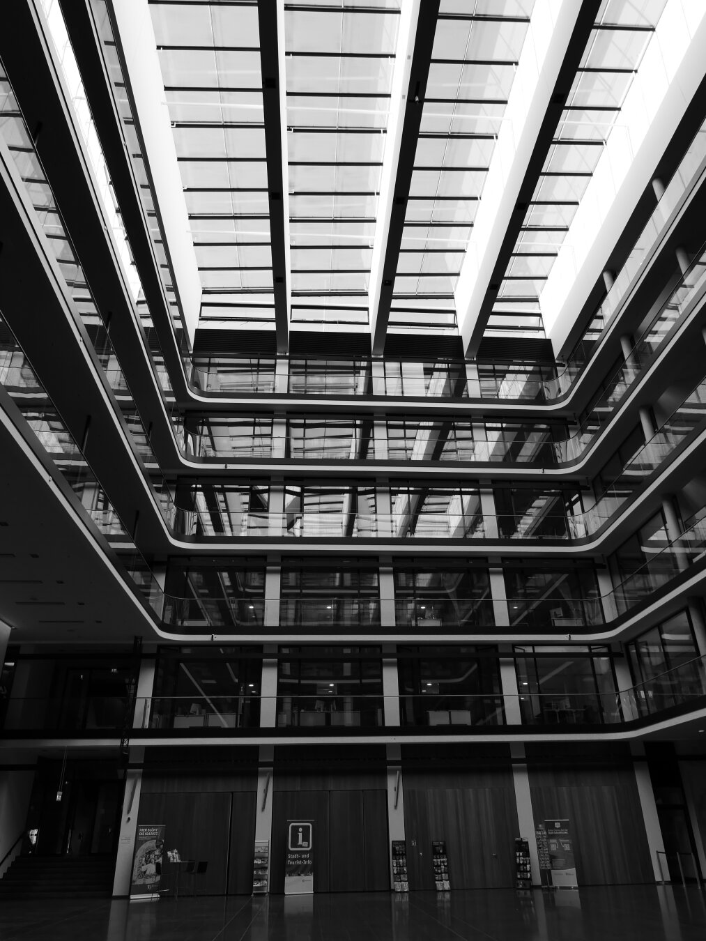 Schwarzweißaufnahme eines modernen, mehrgeschossigen Gebäudeatriums. Der Blick geht von unten steil nach oben zu einem großen Glasdach. Dunkle Stahlträger, Glasflächen und geschwungene Galeriebrüstungen bilden starke geometrische Linien und Kontraste. Im unteren Bildbereich liegt eine weitgehend leere Eingangshalle mit einer Stadt- und Tourist-Information. Die symmetrische Perspektive und das helle Dach verleihen dem Bild eine klare, monumentale Wirkung.
