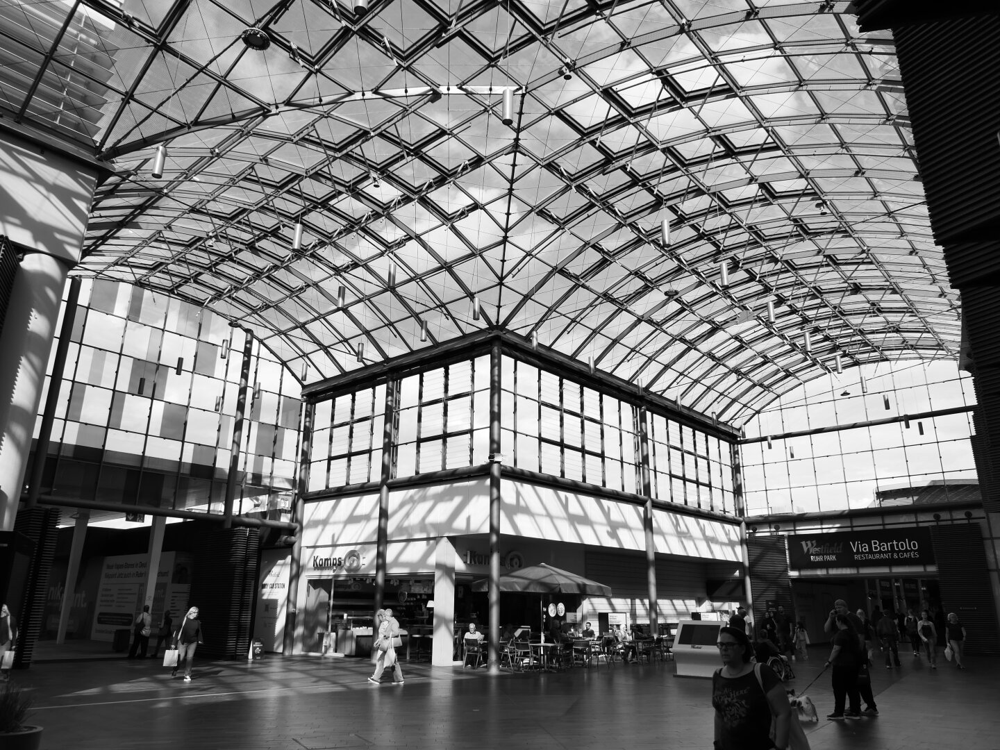 Das Schwarzweißphoto zeigt einen durch ein großes Glasdach überdachten Platz in einem Einkaufszentrum. Zu sehen ist unter dem Glasdach und Glasfenstern, die ca. Zweidrittel des Bildes einnehmen auch eine Bäckerei und einige Menschen.