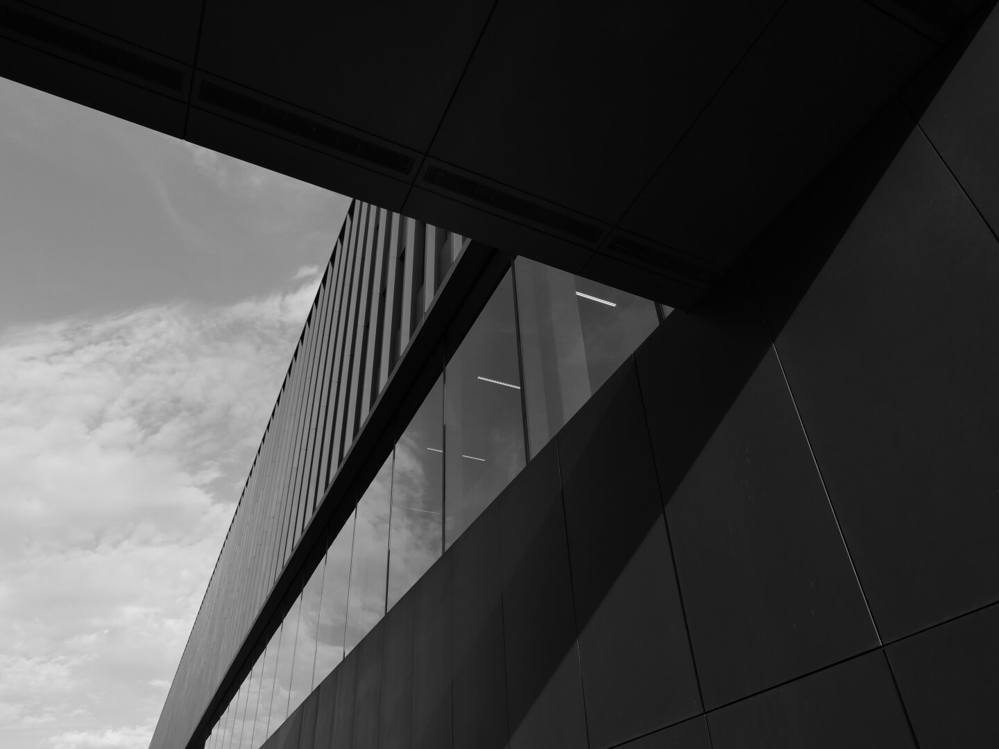 Schwarzweißaufnahme moderner Architektur aus einer stark nach oben gerichteten Perspektive. Dunkle Fassadenflächen, Glasfronten und markante diagonale Linien rahmen einen hellen, teilweise bewölkten Himmel. Die Spiegelungen in den Fenstern und die starken Hell-Dunkel-Kontraste verleihen dem Bild eine abstrakte, grafische Wirkung.