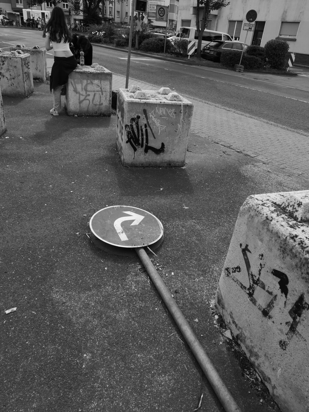 Schwarzweißaufnahme einer städtischen Straßenszene. Im Vordergrund liegt ein umgestürztes Verkehrsschild mit Rechtsabbiegepfeil auf dem Asphalt, umgeben von großen, mit Graffiti besprühten Betonblöcken. Im Hintergrund stehen zwei Personen an einem der Blöcke; dahinter verlaufen eine Straße sowie Wohn- und Geschäftshäuser.