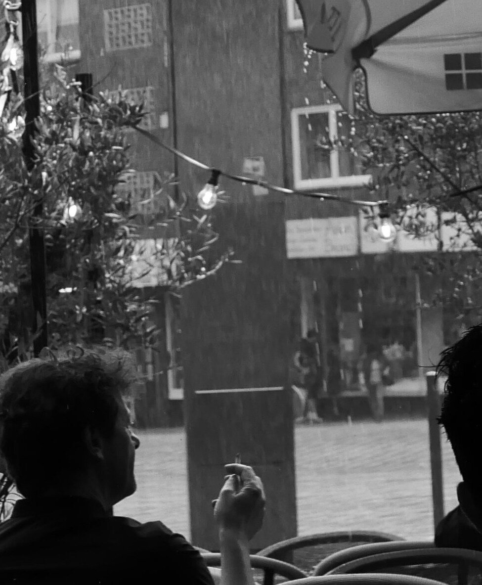 Schwarzweißaufnahme aus einem Café mit Blick durch ein großes Fenster auf starken Regen. Im Vordergrund sitzt eine Person im dunklen Silhouettenprofil und hält die rechte Hand mit einer Zigarette nahe ans Gesicht. Draußen verschwimmen Gebäude und Passanten hinter den Regenschleiern; leuchtende Glühbirnen und Pflanzen rahmen die ruhige, kontrastreiche Szene.