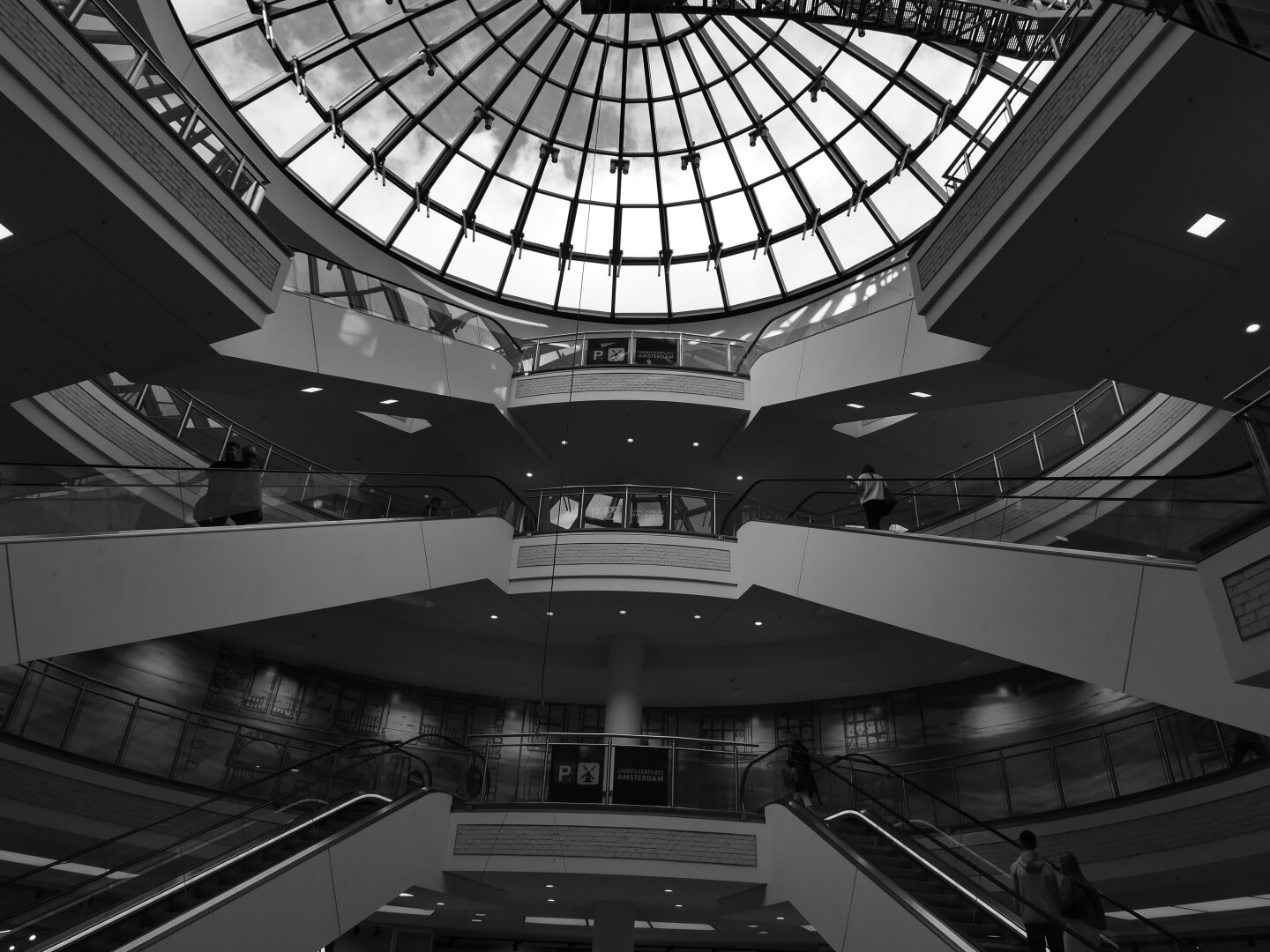 Schwarzweißaufnahme des mehrgeschossigen Innenraums eines Einkaufszentrums. Mehrere Rolltreppen und geschwungene Galerien staffeln sich übereinander und führen den Blick nach oben zu einer großen, runden Glaskuppel. Einzelne Menschen bewegen sich auf den verschiedenen Ebenen und wirken klein innerhalb der weitläufigen, geometrisch geprägten Architektur.