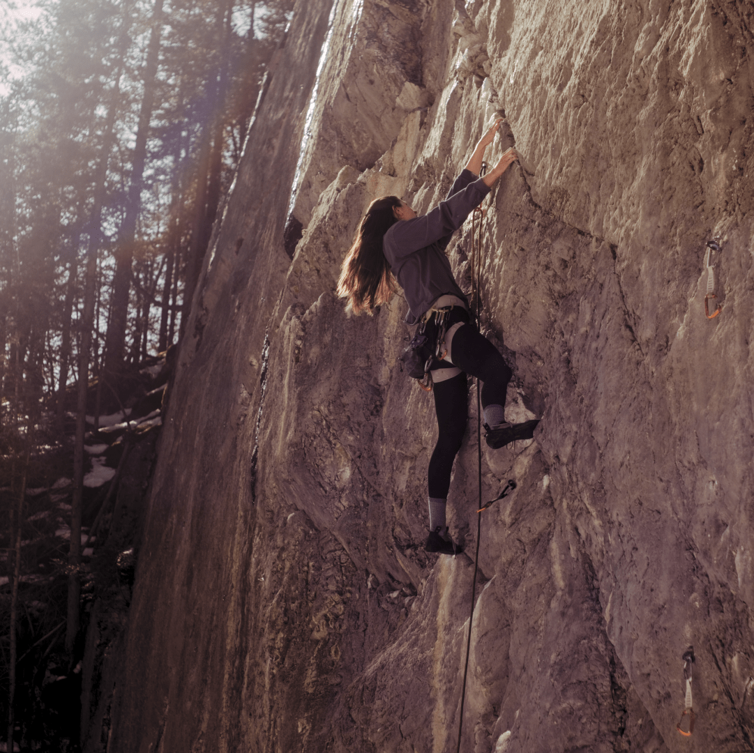 A woman climbs the side of a mountain face in a wintery forest with tools.