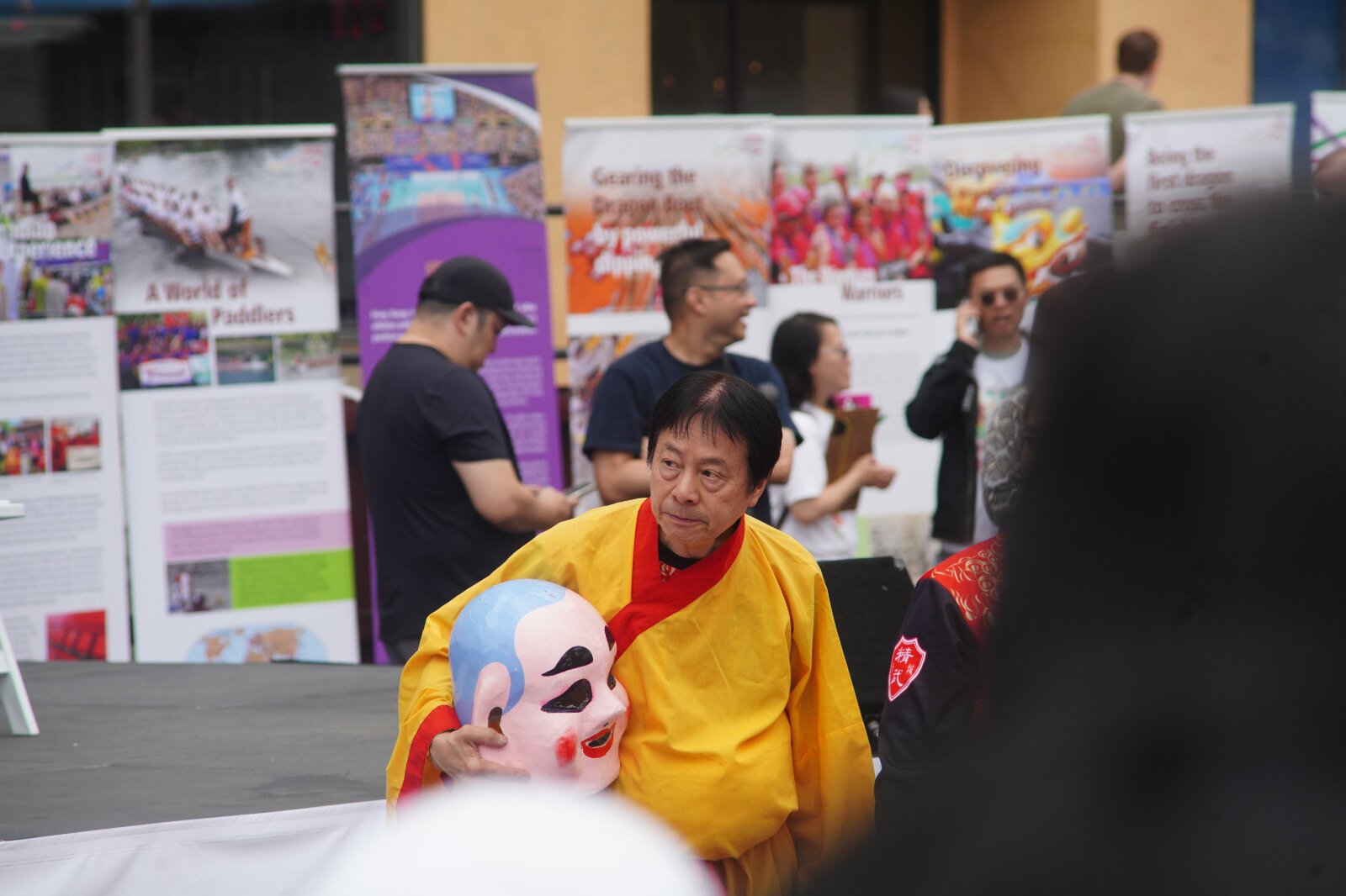 A man in a bright robe holds a mask, while staring glumly into the distance while in front of a crowd.