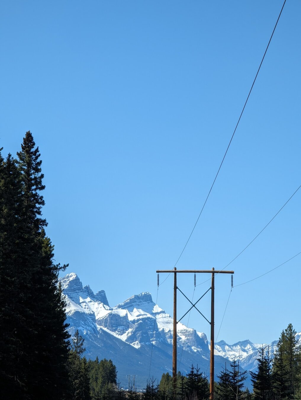 A wilderness scene with dark green trees climbing on the left, in the foreground is a snow capped jagged mountain, in the foreground on the right is a powerline and poles.