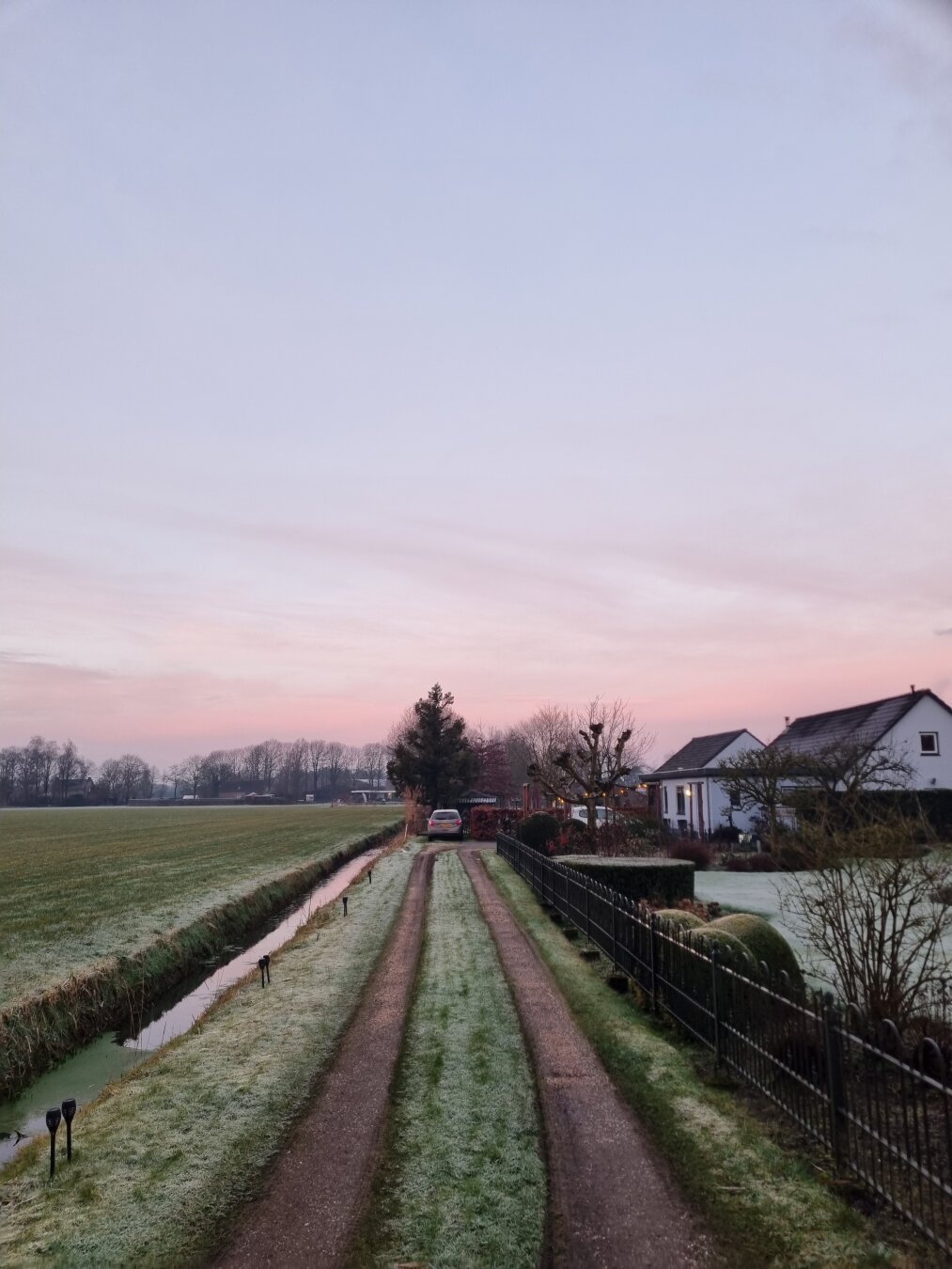 Inrit van gras met stroken van grind voor auto's om op te rijden. Rechts een laag lichtblauw huis. Auto achteraan op de inrit van 50 meter diep.