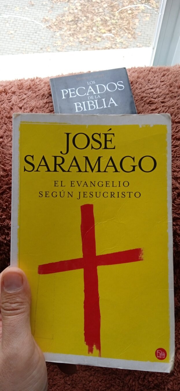 una mano sostiene el libro El evangelio según Jesucristo de José Saramago. La portada del libro es amarilla y un par de trazos rojos en forma de cruz