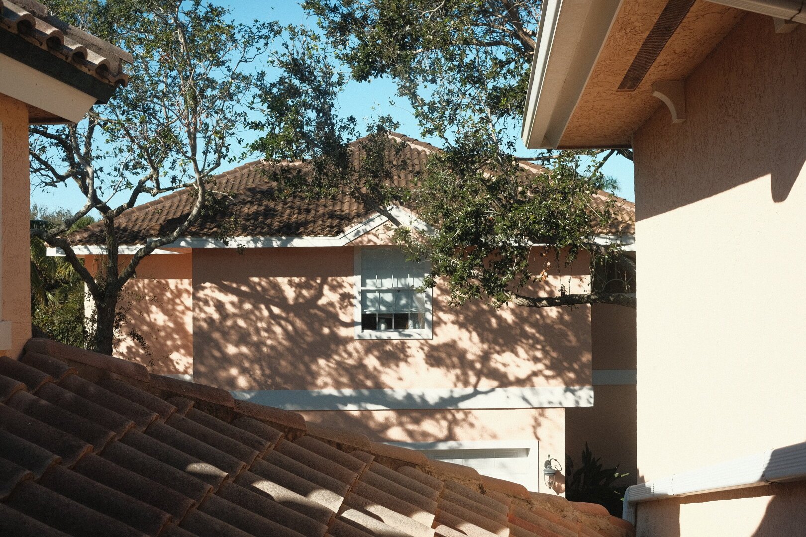 A sunlit residential scene showcasing parts of terracotta-roofed homes with peach-colored walls. A tree with dense foliage casts dappled shadows onto the building's surface. One prominent window on the facade reveals a glimpse of indoor blinds. Architectural details such as eaves and roof tiles add character to the image, and a clear blue sky can be seen above.