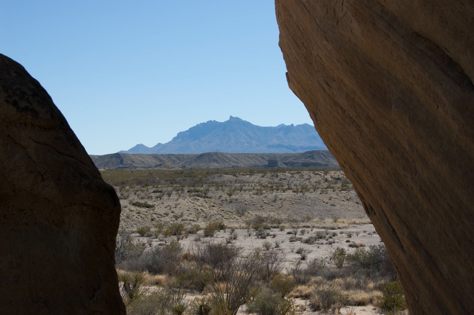 Photograph of the rugged desert terrain of Big Bend National Park in Texas, with a view framed by large rocks, focusing on the distant Chisos Mountains against a clear blue sky.