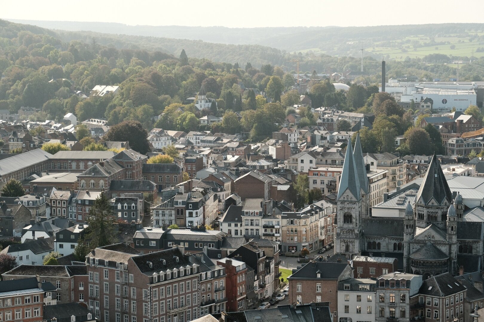 A panoramic view of Spa, Belgium, showcasing densely packed historic buildings with notable church spires. The town nestles amidst verdant hills and trees, with a blend of traditional and modern architecture evident in the landscape.