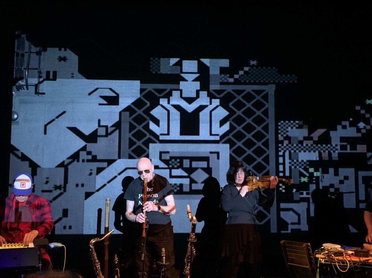 Four musicians playing instruments in front of a video projection of a scene rendered in PETSCII in grey