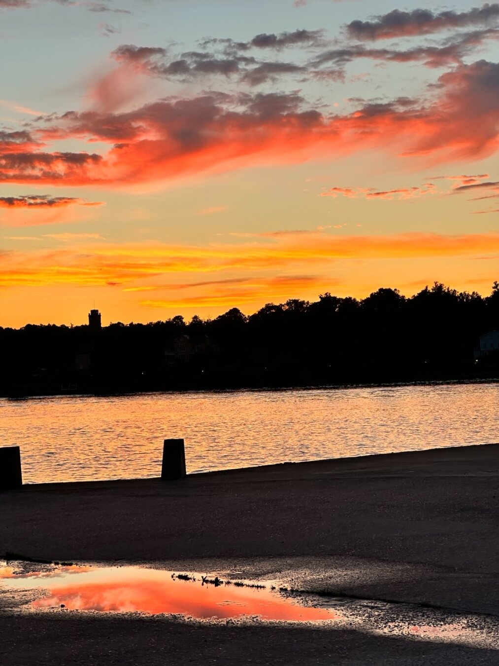 an orange-pink cloud in the sunset sky reflecting in a puddle on dark ground. Beyond that is a body of water lit with radiant orange light and the silhouette of the distant shore