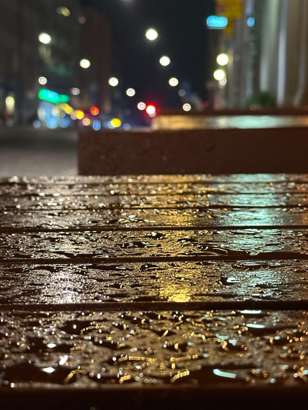The surface of an outdoor table covered with raindrops and reflecting white, amber and green lights. In the distance is a blurred night-time urban streetscape.