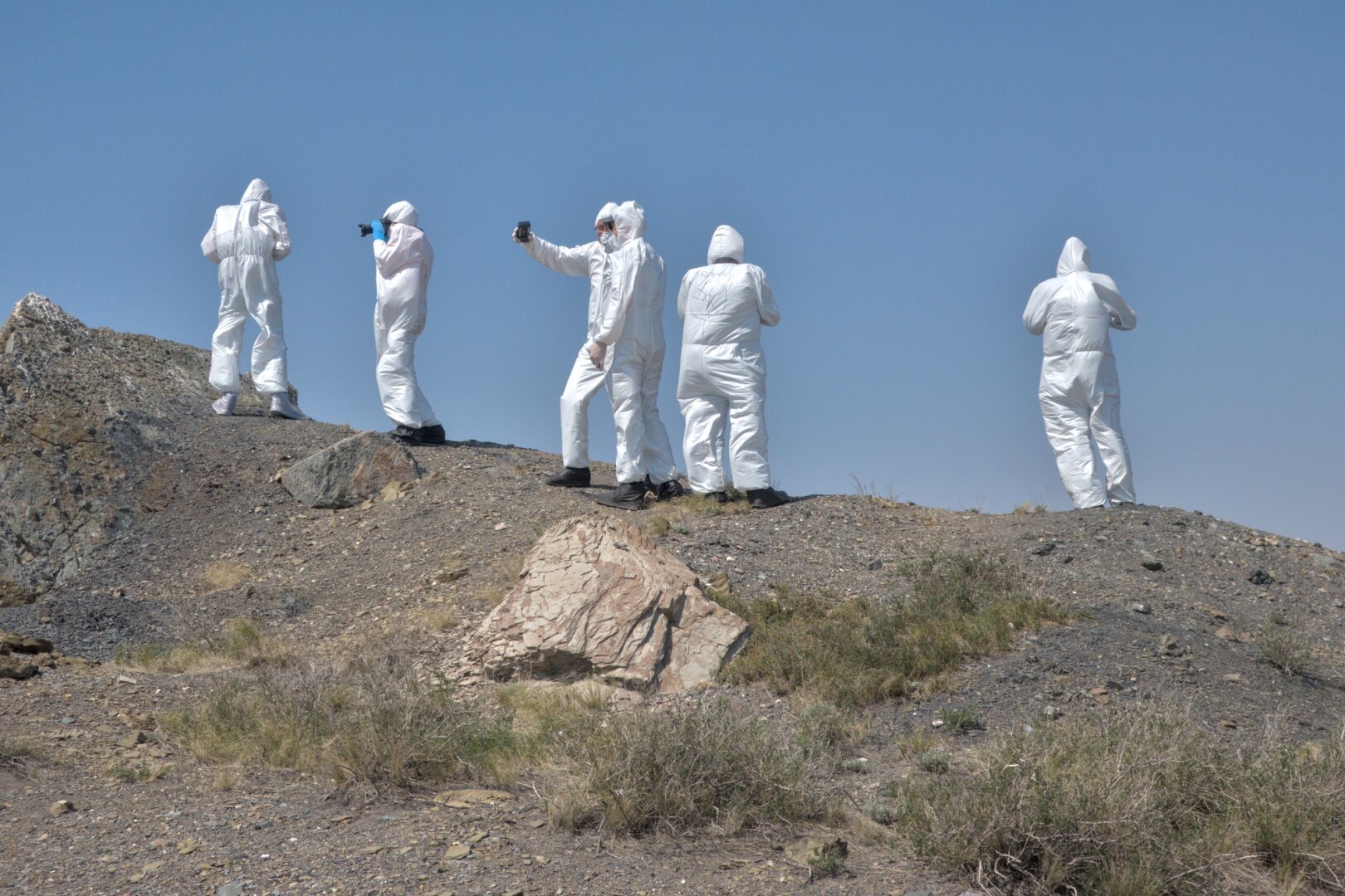 A group of people in HAZMAT suits stand on a a heap of earth. One holds a mobile phone ready to take a selfie.