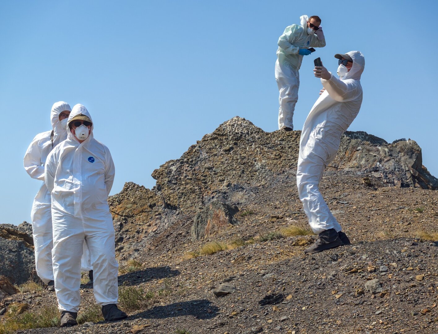 A group of people in HAZMAT suits stand on a a heap of earth. One holds a mobile phone ready to take a selfie.