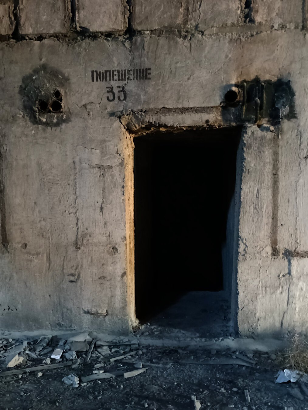 A concrete wall with a doorway leading into darkness. There is Cyrillic text stencilled on the wall. Rubble is strewn across the floor