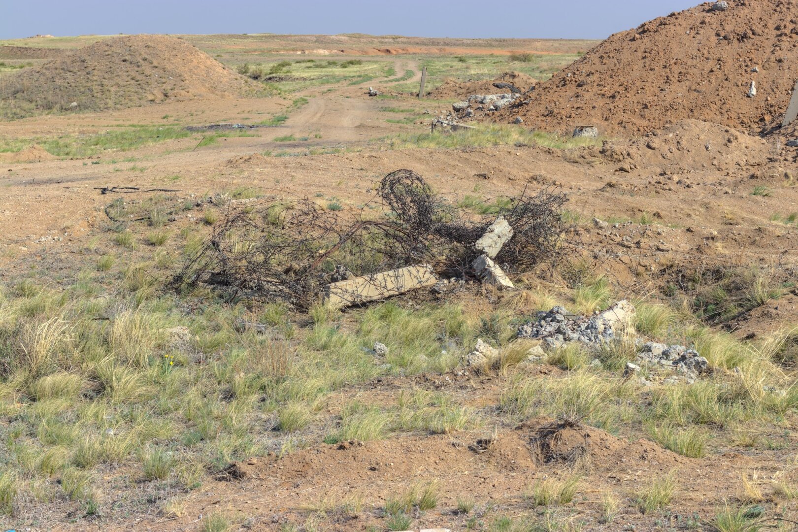 A dirt road winds it's way through the wreckage. There are mounds of earth on both sides and a pile of concrete and barbed wire in the centre of the frame.