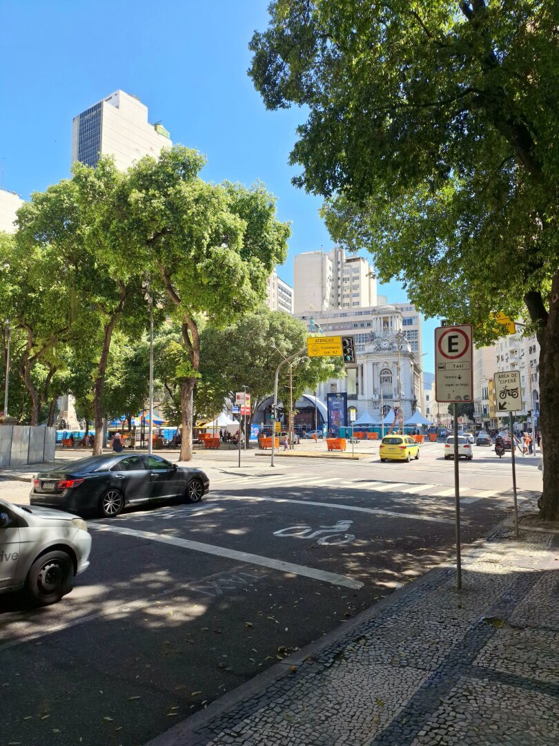A bright daytime scene in Cinelândia, Rio de Janeiro during pre-carnival, showing a wide street corner with tall leafy trees, mosaic sidewalks, and passing cars. Modern high-rises and an ornate historic building stand in the background, adorned with banners for the upcoming festivities.
