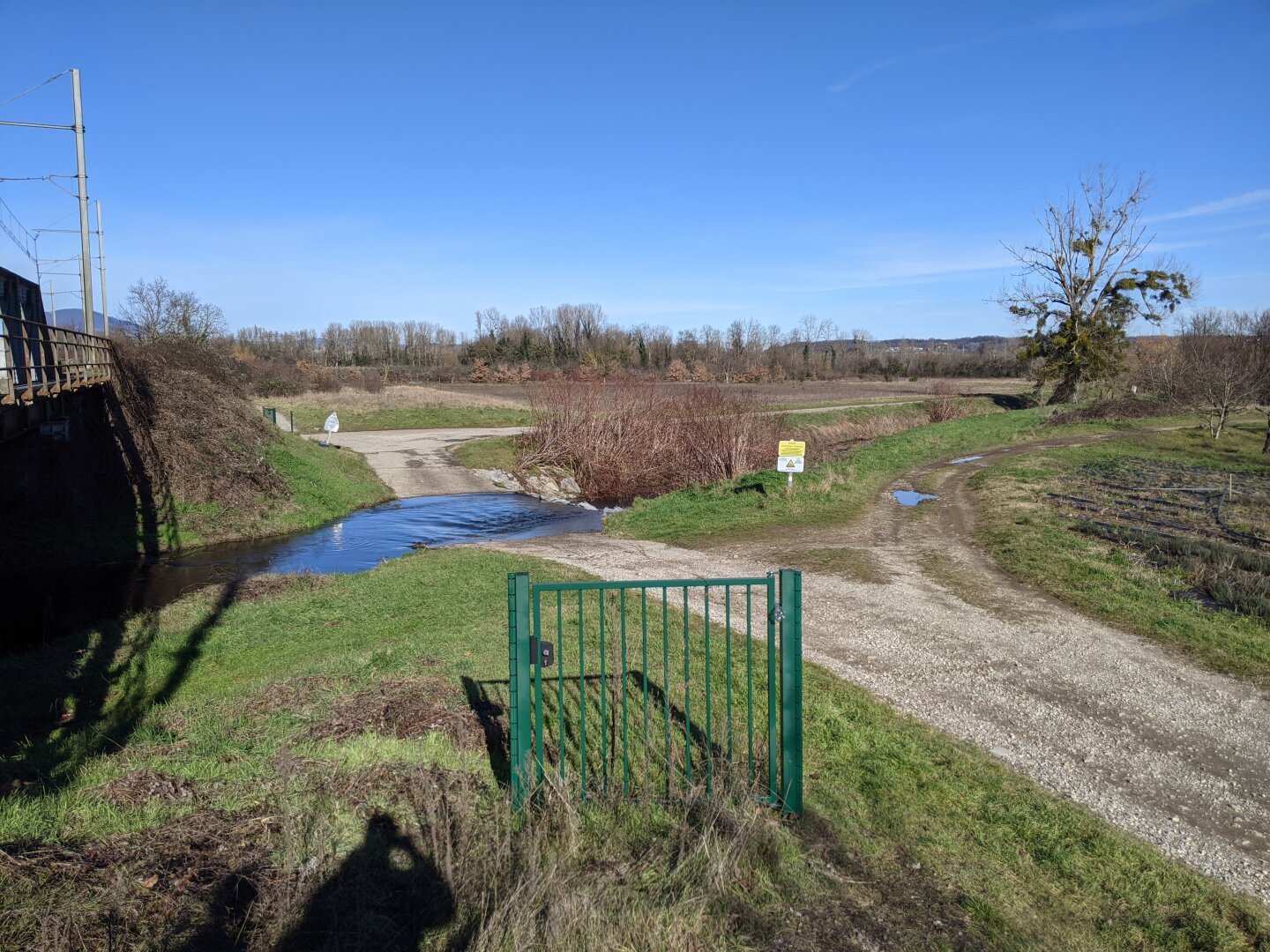 A sunny winter country landscape with a dirt road and a small river in the background.
In the foreground, there is a solitary gate (about 1m high), no other barrier to extend it. There never was (no remain visible in the grass).
It is locked but you can go around it.