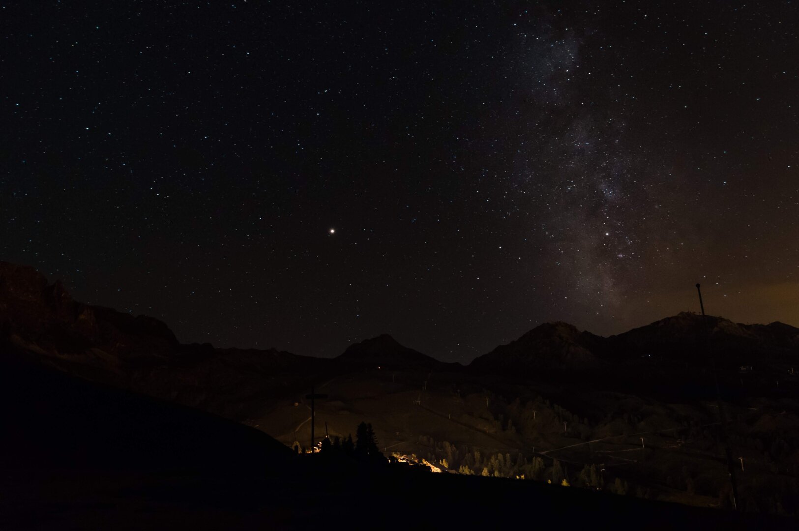 Photo taken during the night in summer. Wide angle. There are mountains in the foreground and the milky way in the background.
Lights in the middle coming from the ski resort partially hidden by the mount.