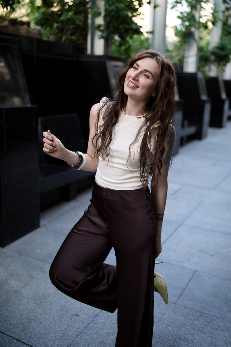 A fashion like portrait, where a young Ukrainian woman is standing on one leg, showing her high heel. She has long loose hairs, and wearing a modern outfit, white and dark plumb.
