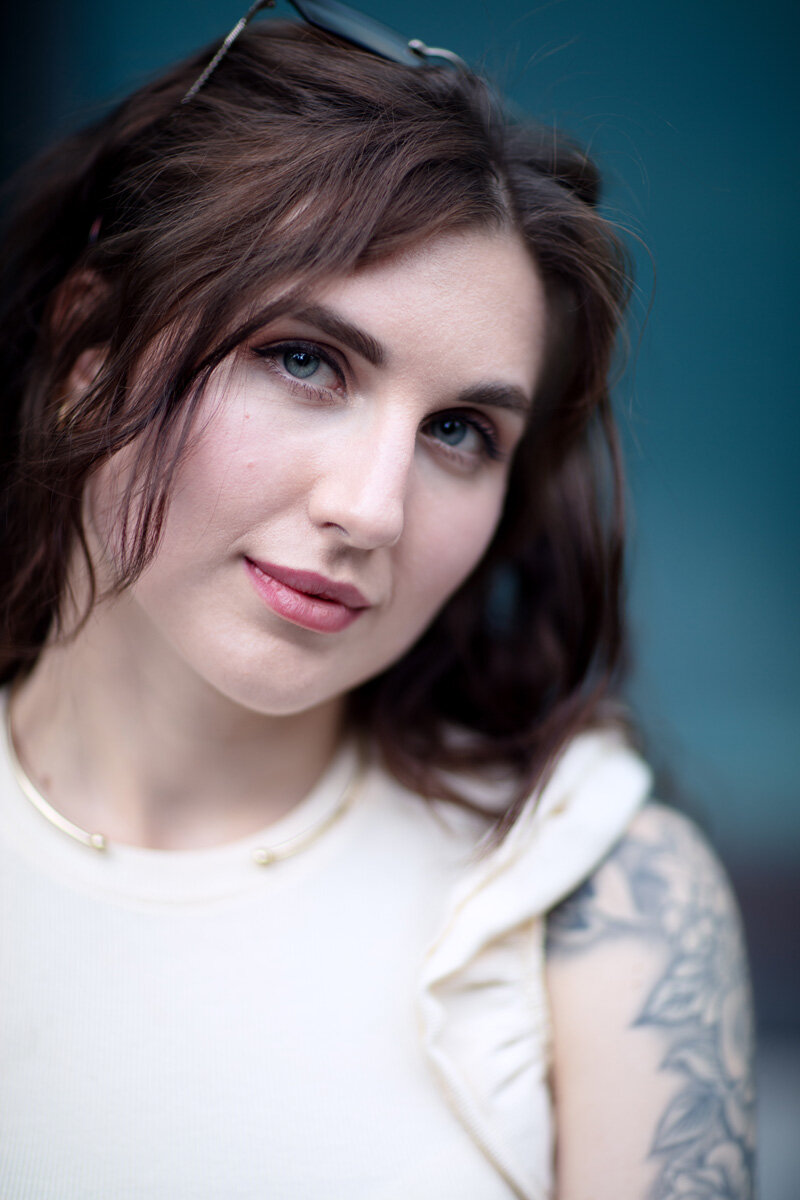 A close up portrait of a young Ukrainian woman. Her light skin tone with her white outfit contrasts with her blue eyes and blue blurry background. She is looking strait to the camera, with a subtle smile.