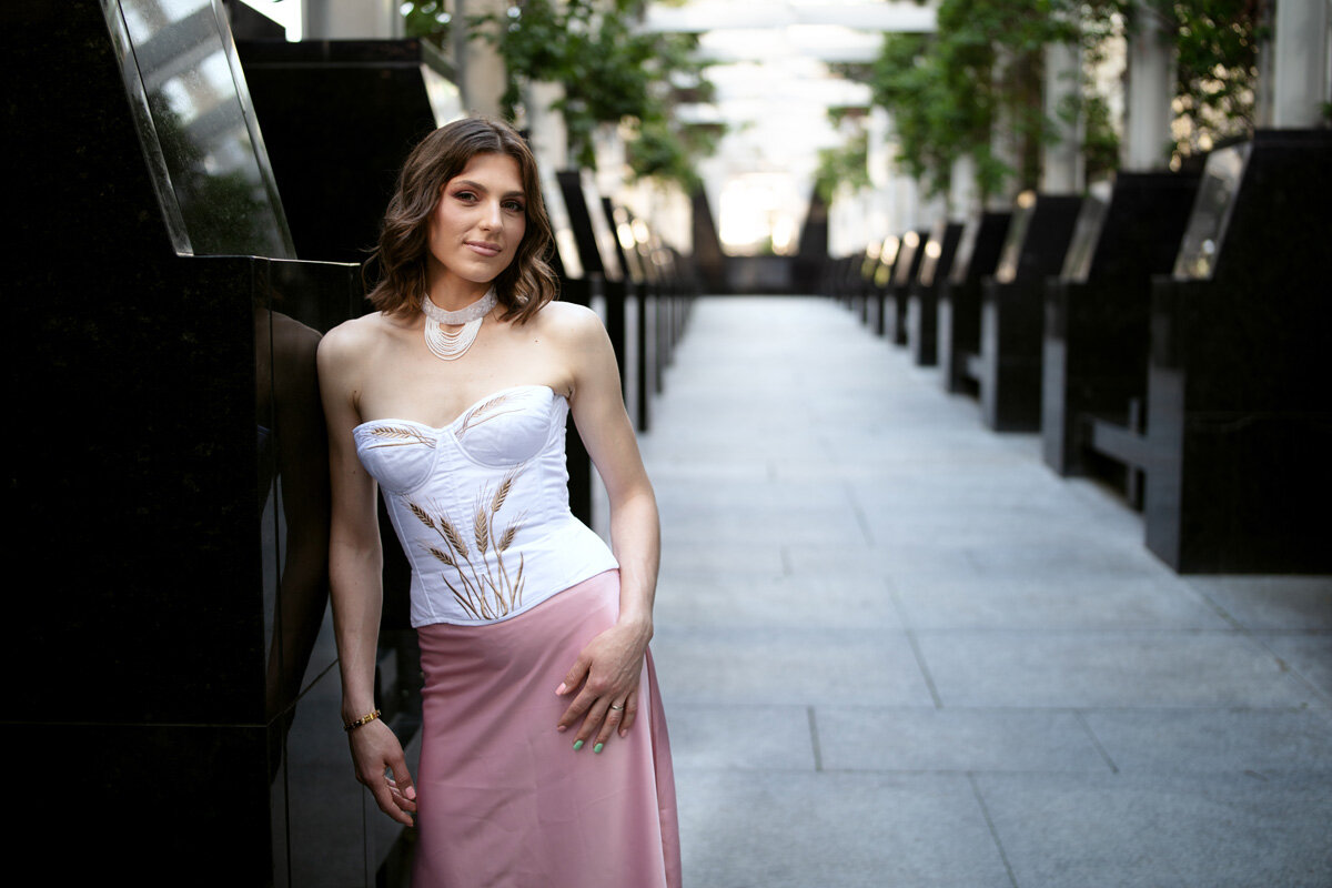 A fashion like portrait, a young Ukrainian woman is standing in a architectural alley with black stone's borders and white floor. There is some vegetation that adds some green to the image. The model is wearing a traditional Ukrainian outfit, the top is white without sleeves and with wheat iconography, the skirt is pale pink. She is smiling slightly.