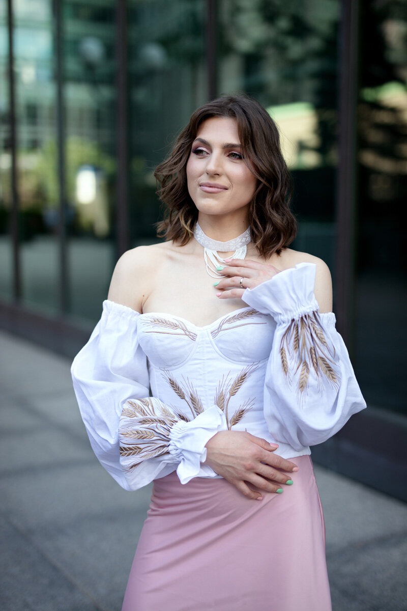 A portrait of a young Ukrainian woman, wearing a traditional Ukrainian outfit. The outfit is white and light pink displaying wheat related iconography.