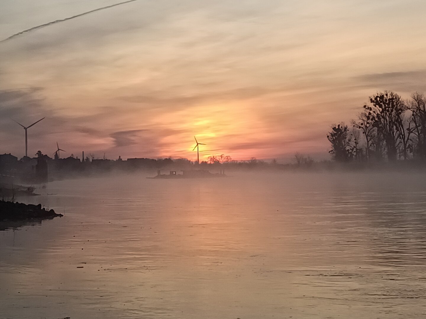 wir blicken zum Sonnenaufgang auf die Elbe.
im Osten färbt die Sonne die Wolken rötlich gelb und über dem Fluss steht eine dünne Nebelschicht.
Über den Fluss fährt im Nebel, eine Fähre von Ufer zu Ufer.

Die Sonne geht genau hinter einem Windrad auf.