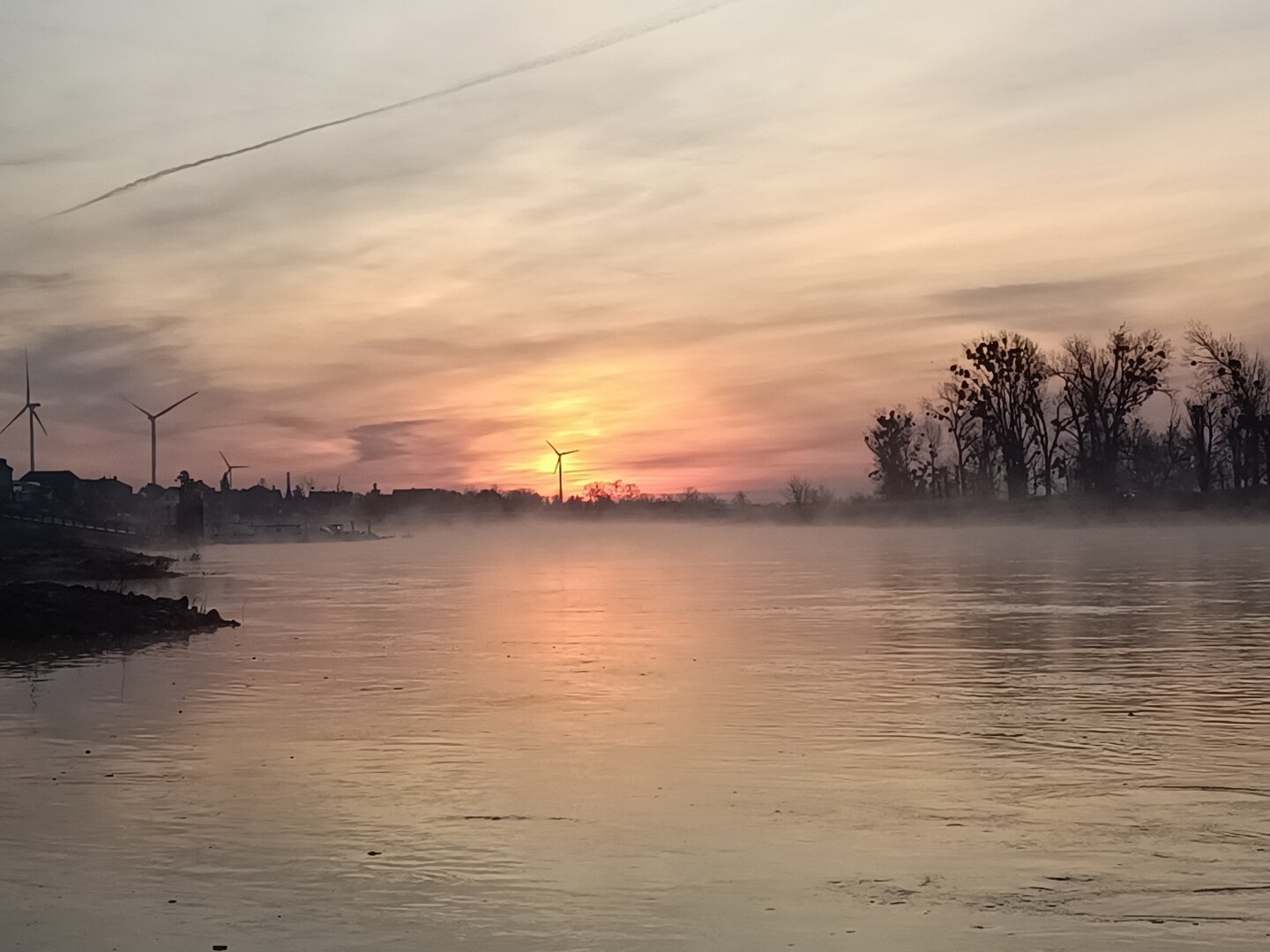 wir blicken zum Sonnenaufgang auf die Elbe.
im Osten färbt die Sonne die Wolken rötlich gelb und über dem Fluss steht eine dünne Nebelschicht.
Über den Fluss fährt im Nebel, eine Fähre von Ufer zu Ufer.

Die Sonne geht genau hinter einem Windrad auf.