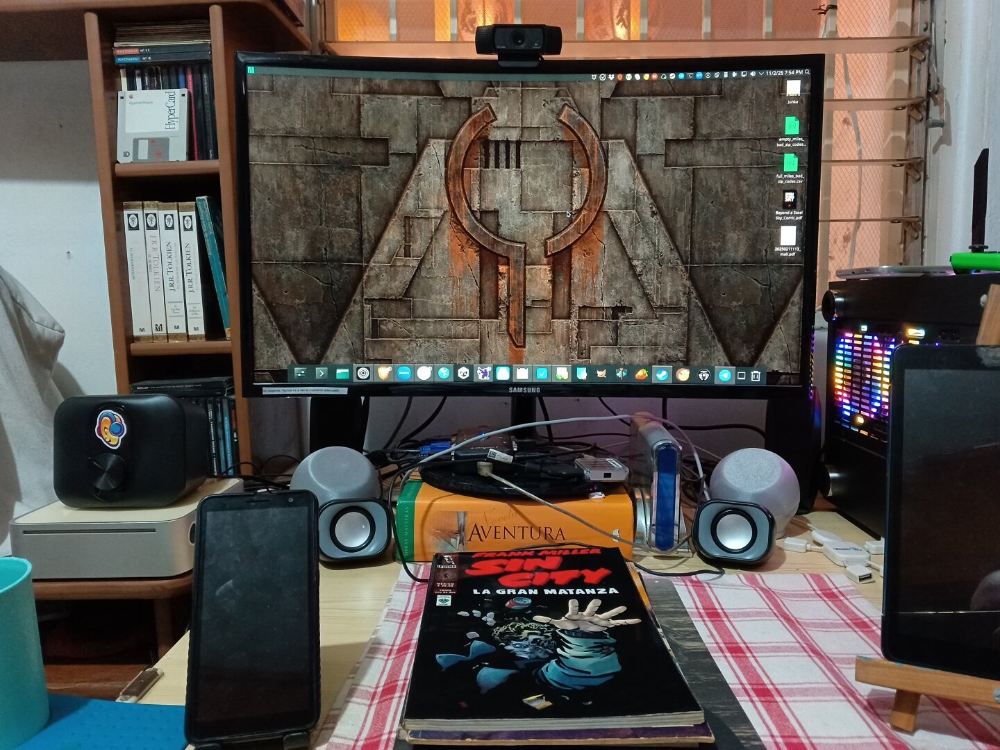 A photo of a cruved LCD monitor showing a KDE desktop with a Quake wallpaper. A Sin City comic book is in front of the monitor.