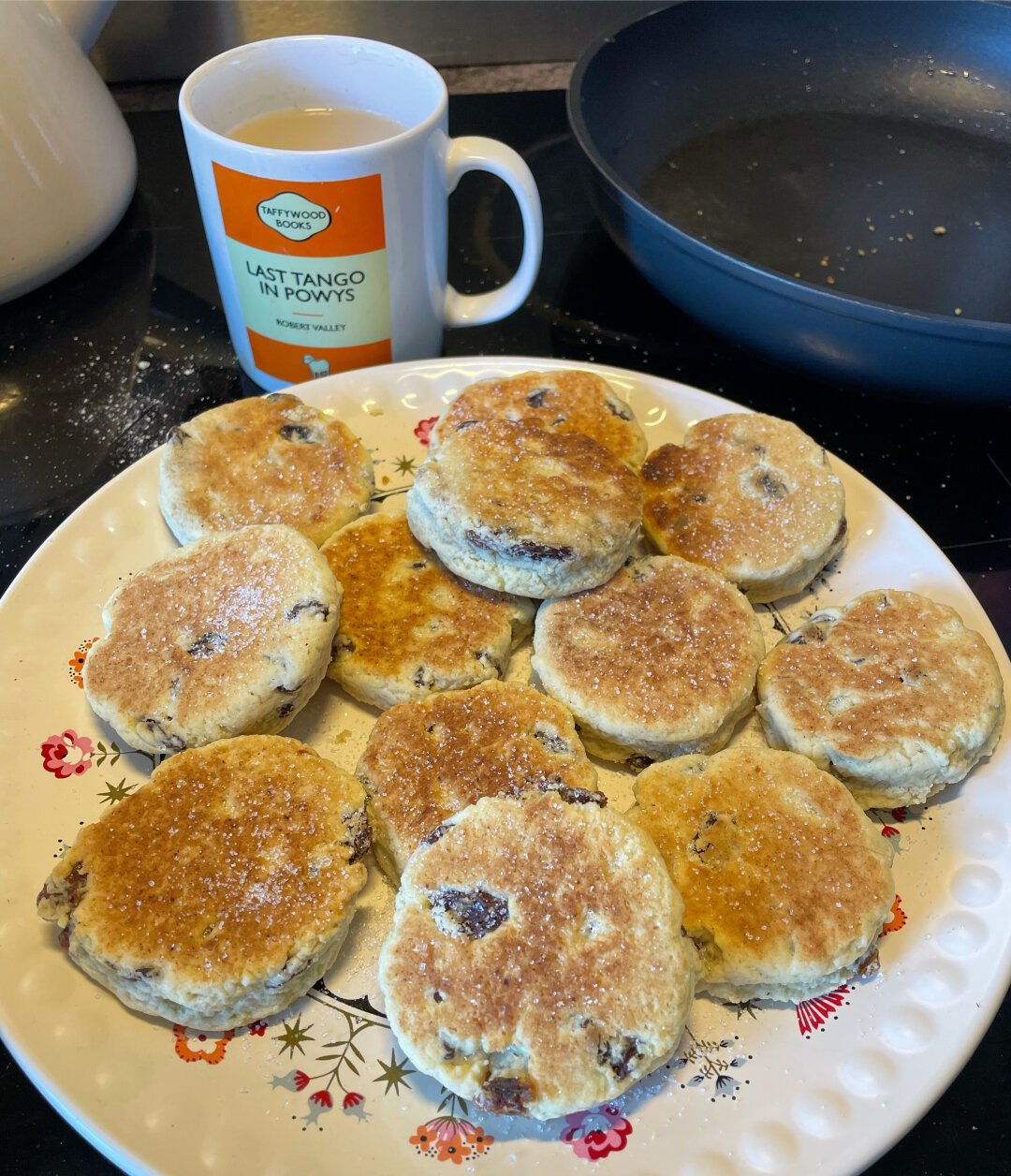 A plate of homemade Welsh cakes with a mug behind the plate. The mug is in the style of an old Penguin paperback book cover, with the title, Last Tango in Powys.