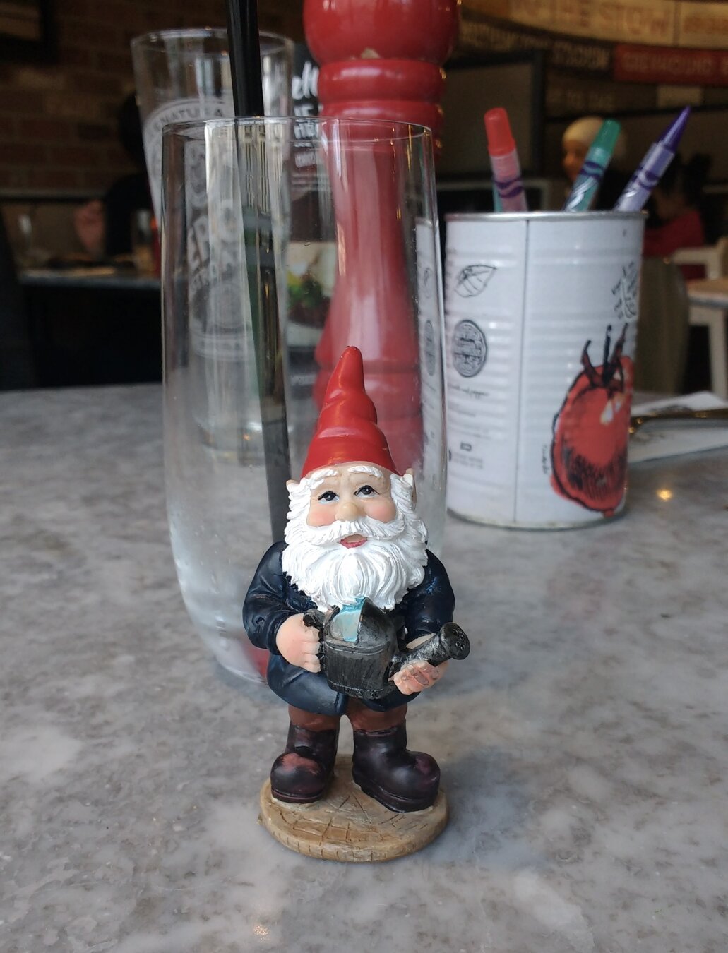 A small, resin gnome figure holding a watering can, stands front and centre on a table in Pizza Express. Behind it, empty glasses, a red peppermill and a tin of crayons.