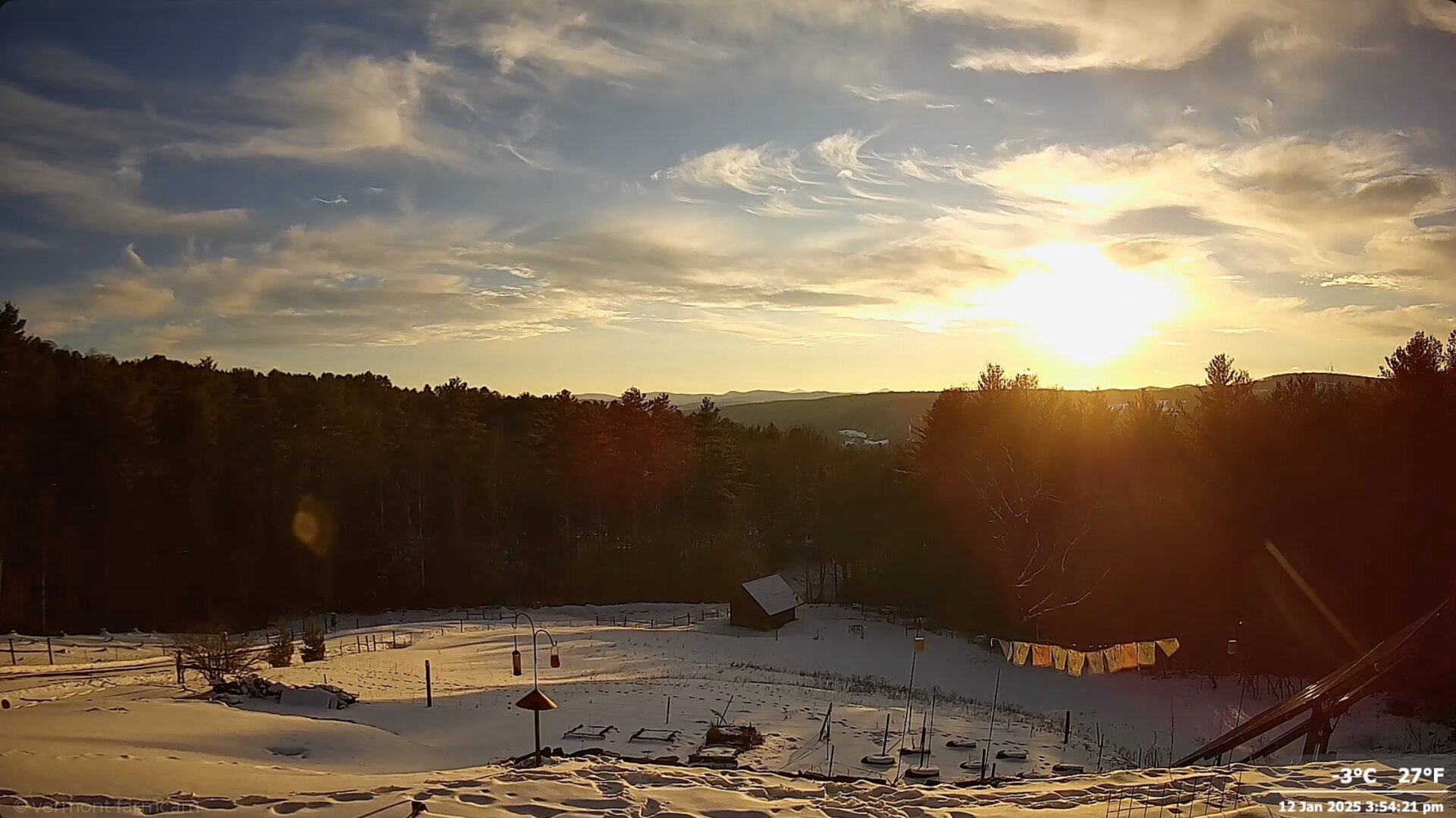 the sun is setting behind clouds and mountains coloring wispy clouds in yellows and golds. The sun is reflecting on the snow in the foreground.