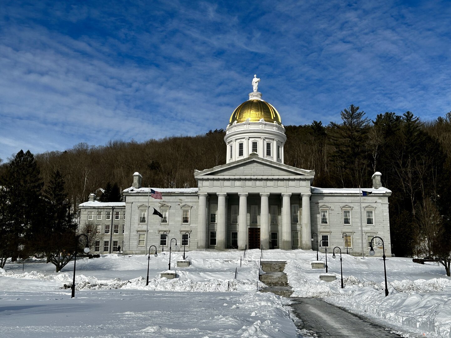 Montpelier’s capital building’s golden dome shining in the afternoon winter sunshine. With a bright blue sky and trees behind the building.