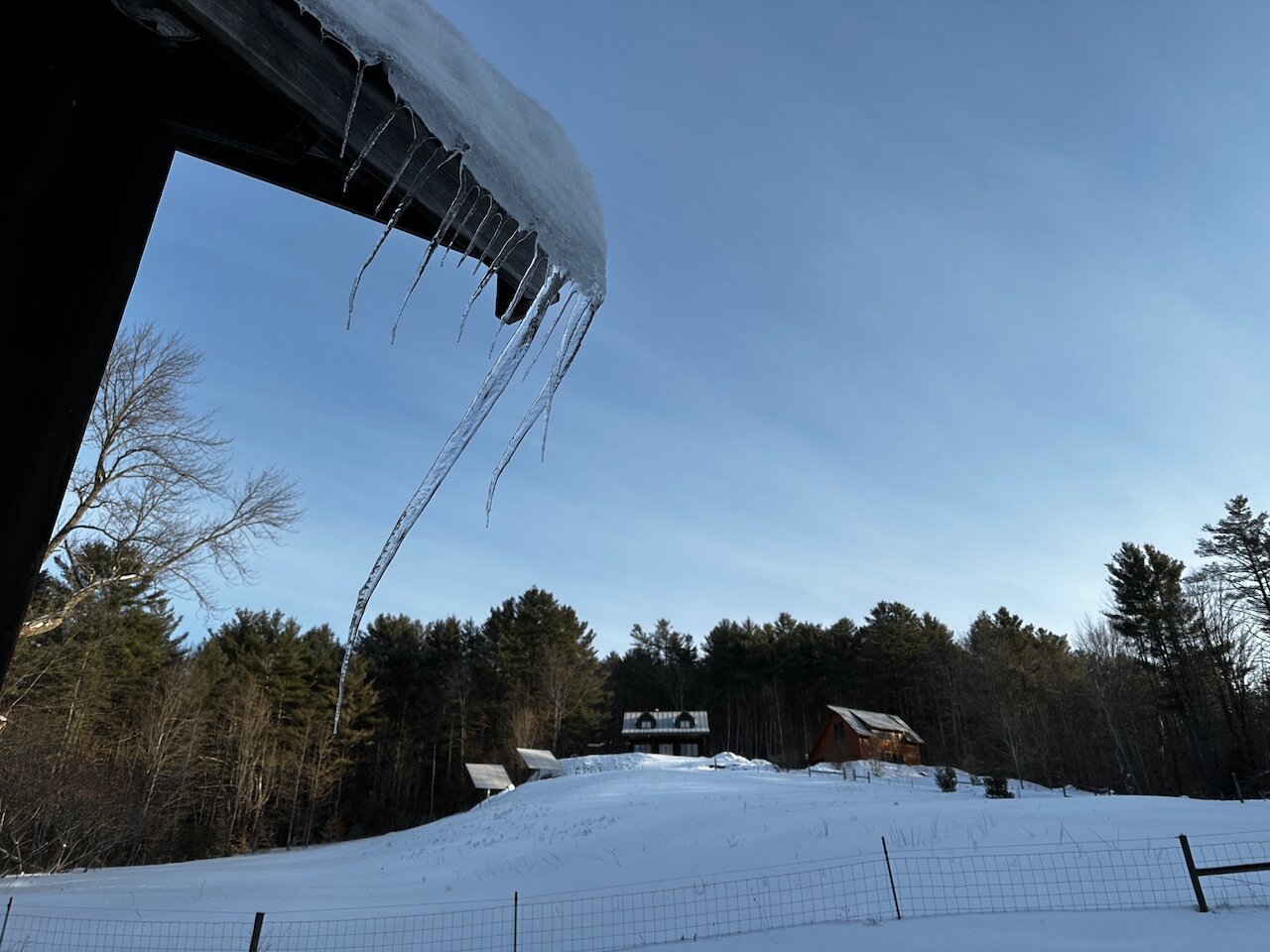 In the foreground jagged icicles hang at an unusual angle due to wind. In the background a house, some solar panels and another building can be seen past at the snowy fields.