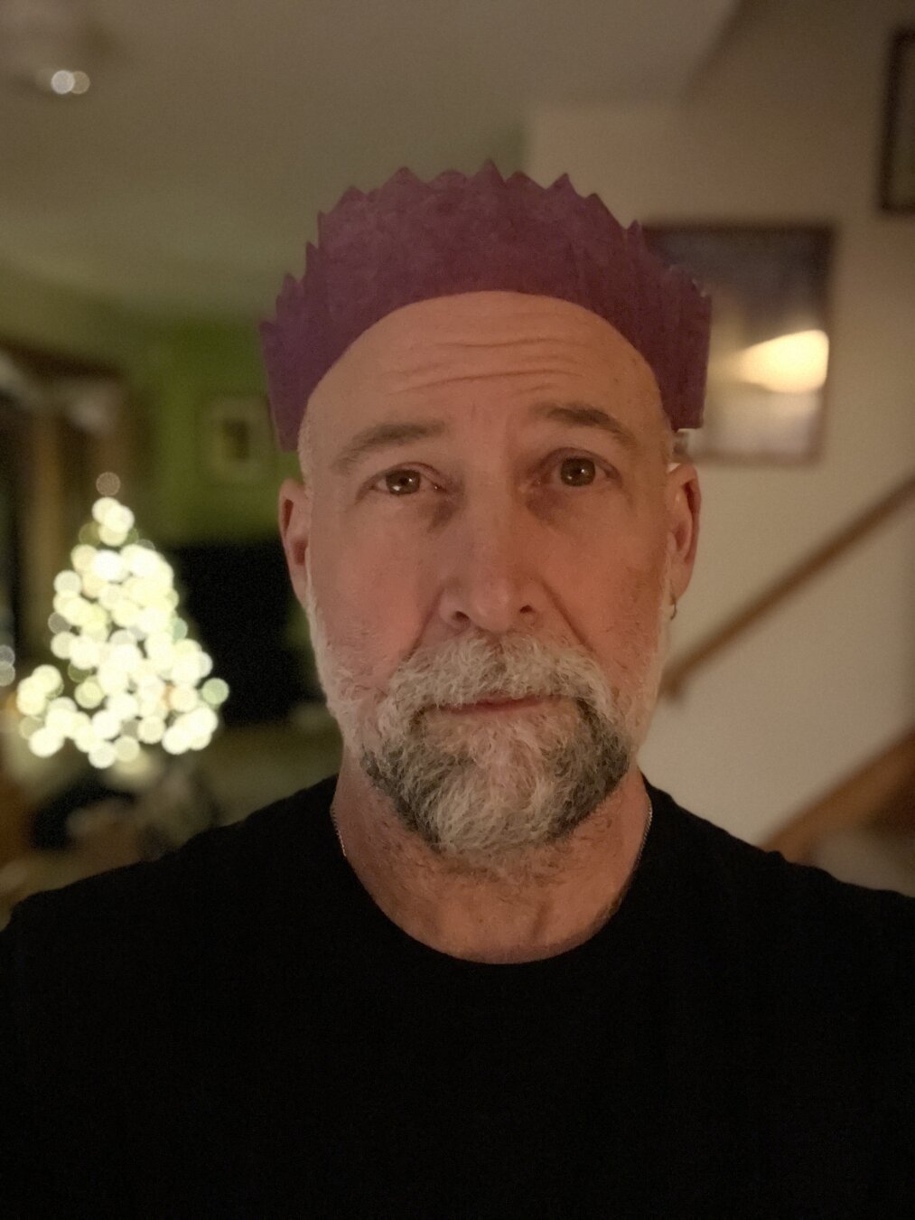 An older white man with a salt and pepper beard — and a slightly creepy uncle vibe — is looking directly at the camera and is wearing a purple paper crown from a Christmas cracker. An out-of-focus Christmas tree with all white lights is illuminated in the background.