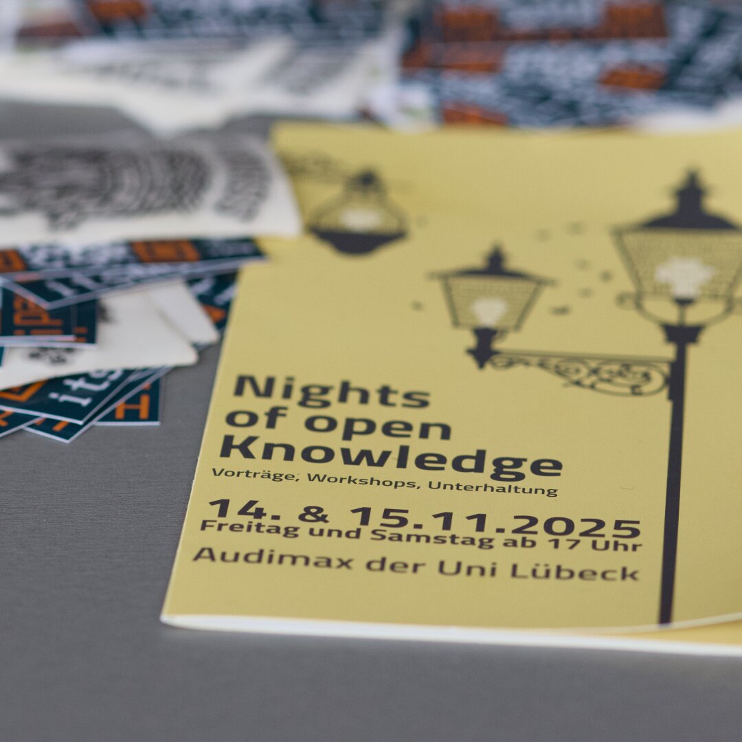 Ein gelbes Flyer-Cover mit der Aufschrift „Nights of open Knowledge – Vorträge, Workshops, Unterhaltung – 14. & 15.11.2025 Freitag und Samstag ab 17 Uhr – Audimax der Uni Lübeck“. Daneben liegen unscharfe Sticker. Auf dem Flyer sind drei stilisierte Straßenlaternen zu sehen.