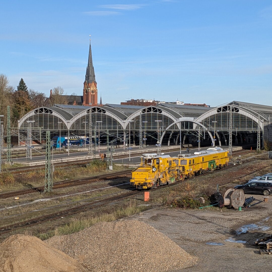 Bahnhofsszene an einem sonnigen Tag: Vor den bogenförmigen Glasdächern eines Kopfbahnhofs steht ein leuchtend gelber Gleisbauzug auf einem Nebengleis, im Hintergrund fahren blaue Züge und links ragt der Turm einer Kirche über die Gebäude.