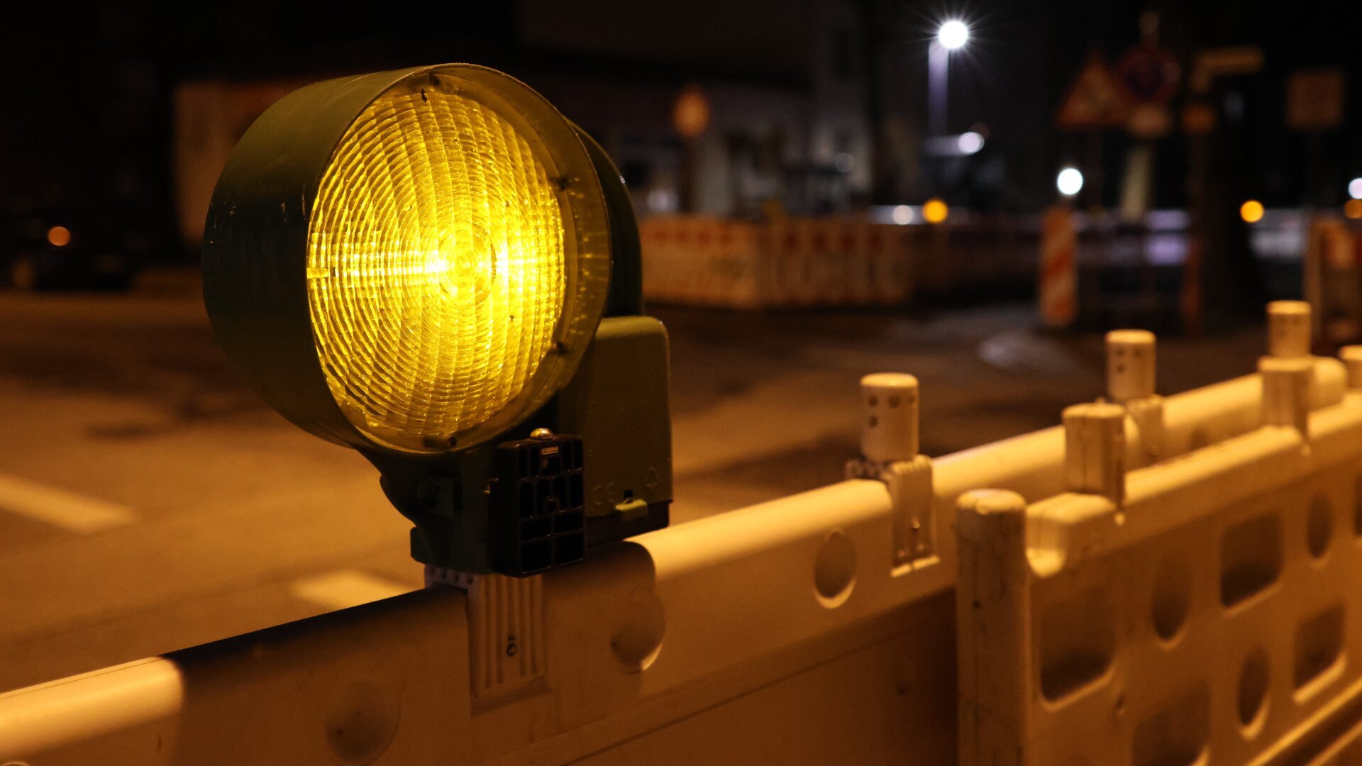 Gelbe Baustellenleuchte auf einer rot-weißen Absperrung einer Baustelle. Nacht. Im Hintergrund Lichter verschiedener Straßenlaternen.