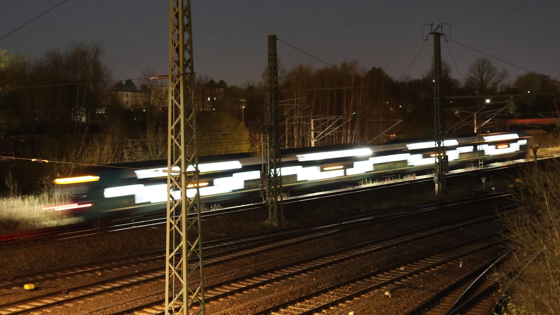 Gleise bei Nacht. Ein vorbei fahrender Personenzug ist stark beleuchtet und hat viel Bewegungsunschärfe.