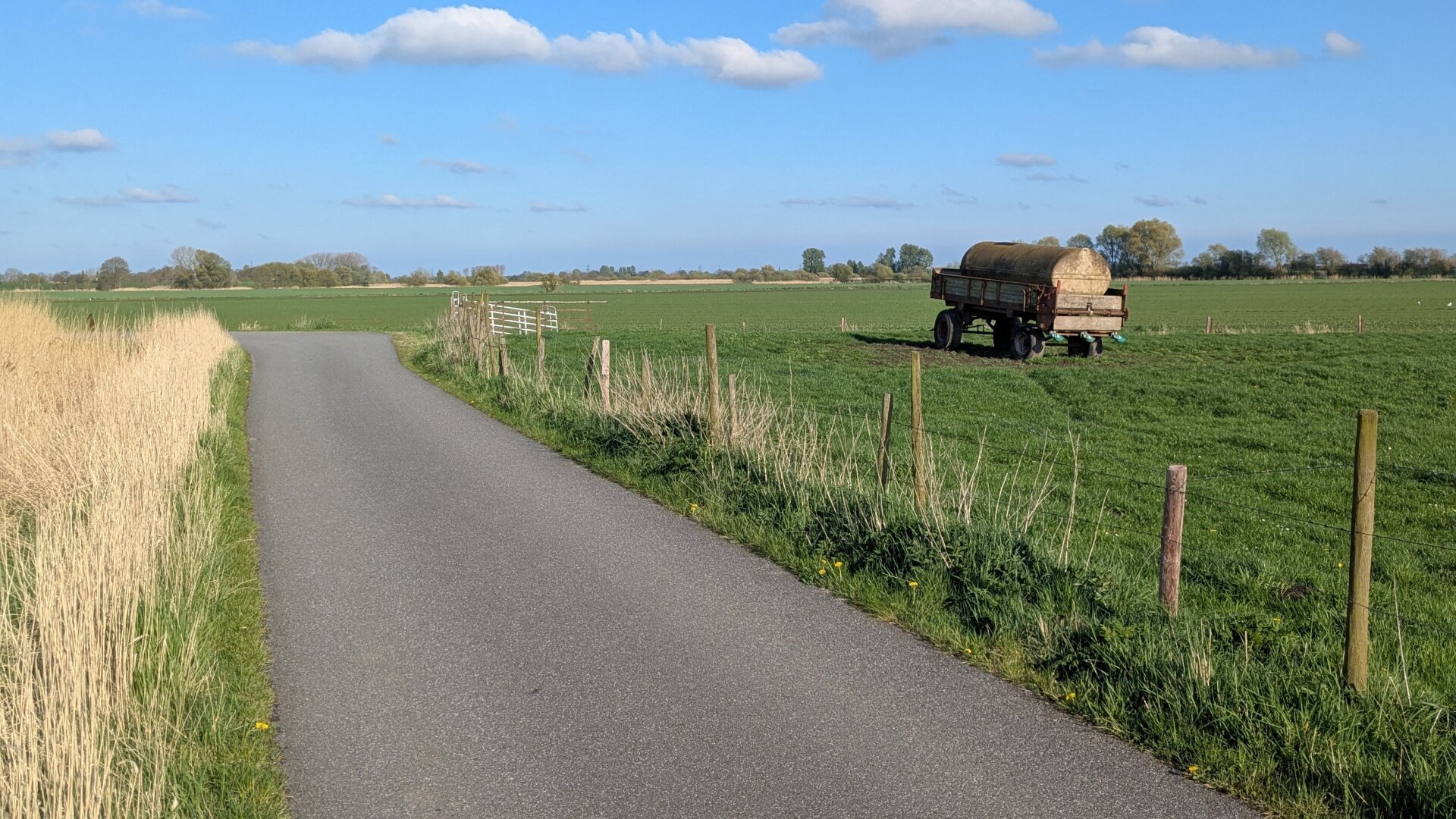 Eine schmale Straße führt geradeaus und biegt am dann links ab. Rechts der Straße ist ein Zaun und dahinter eine grüne Wiese. Auf der Wiese steht ein landwirtschaftlicher Anhänger mit einer Viehtränke. Die Sonne scheint, der Himmel ist blau mit einigen weißen Wölkchen.