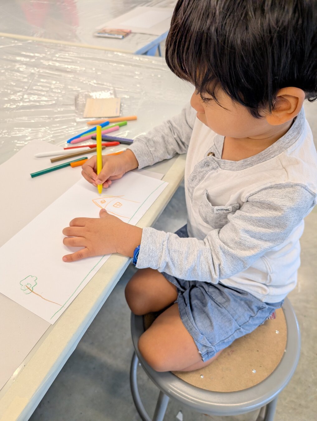 Young child sitting at a table drawing with crayons. He's wearing a grey long sleeve shirt and blue shorts.