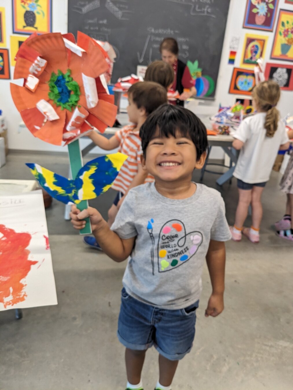 Young boy in a grey t-shirt and jean shorts holding an orange flower made of paper