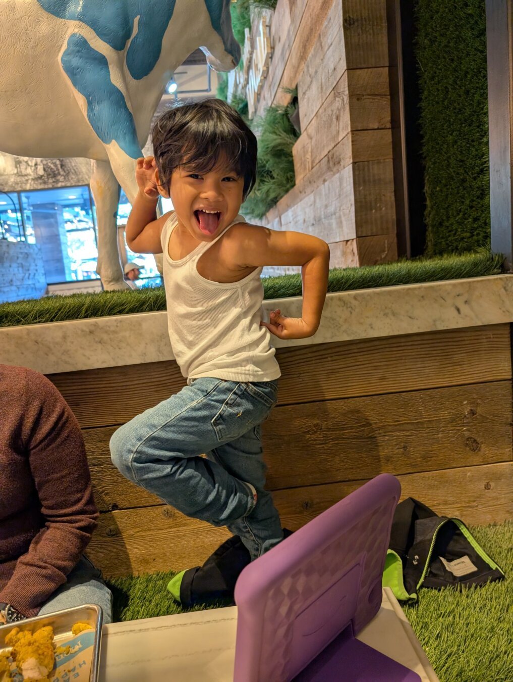 A young boy in a white tank top and jeans poses playfully, sticking his tongue out, with a cow sculpture and wooden wall behind him. A portion of another person in a burgundy sweater, a purple tablet, a plate of food, and a black jacket are also visible in the image.