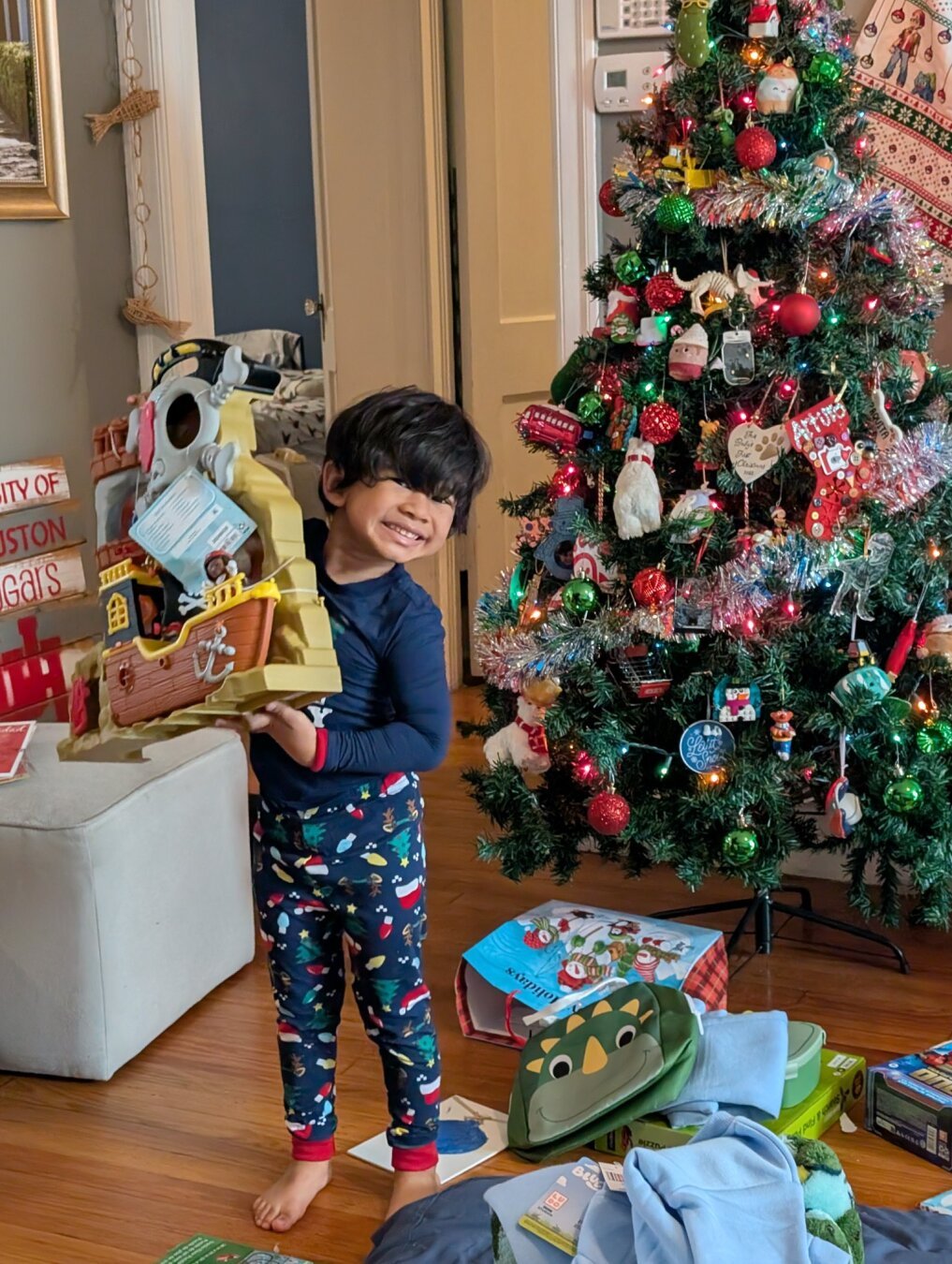 A young child stands smiling while holding a yellow and brown toy pirate ship in front of a decorated Christmas tree. The child is wearing navy blue Christmas pajamas with a festive pattern and red cuffs. The living room setting includes hardwood floors, a white ottoman, and what appears to be a 