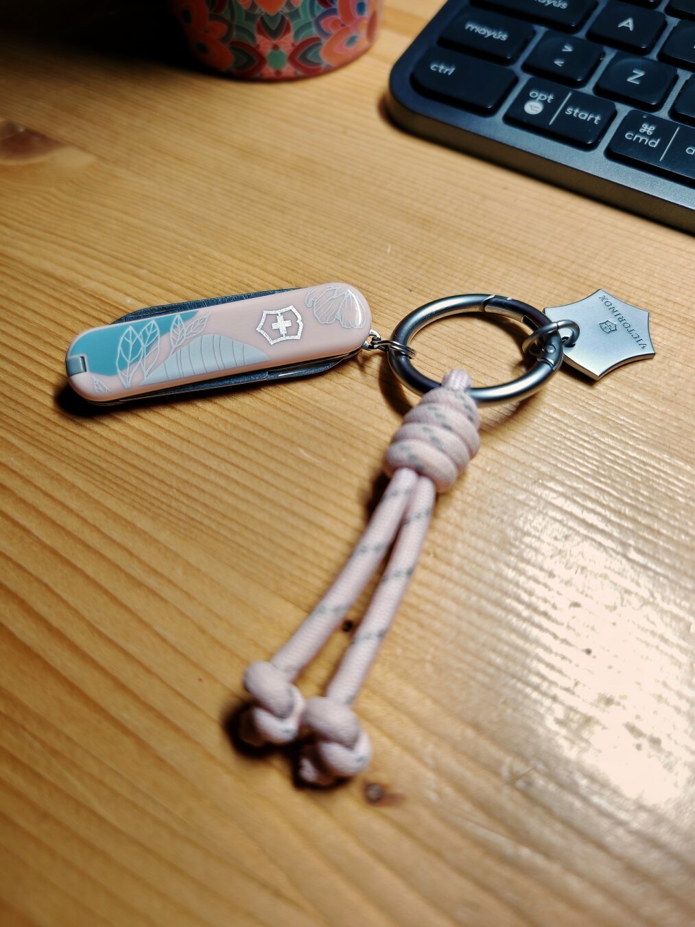 Pequeña Victorinox de 58mm (classic sd) con cachas en rosa y azul pastel, con un pequeño dibujo blanco y el escudo de Victorinox (corresponden a la versión París Style). De su anilla cuelga un llavero con un trocito de paracord rosa con pintas grises y un escudo de metal de la marca. Todo ello delante de un teclado y el fondo de una taza y sobre una mesa de pino.