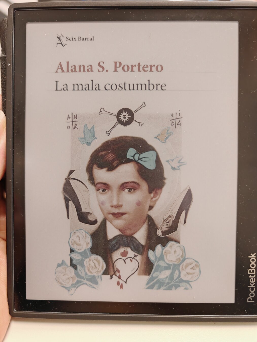 En la pantalla de un eReader a color se ve la portada del libro «La mala costumbre» de Alana S. Portero publicado por Seix Barral. Diseñada por Roberta Marrero, representa la estampita de Santo Domingo Savio, alumno de San Juan Bosco, con algo de maquillaje, un lazo y rodeado de un par de tacones, unos grafitis y unas flores