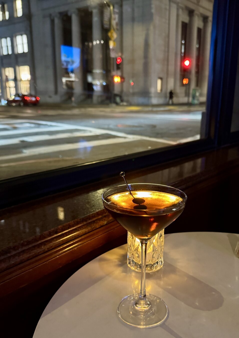 Picture of a Manhattan cocktail on a table with a candle behind it - view is a financial district intersection in San Francisco at night.