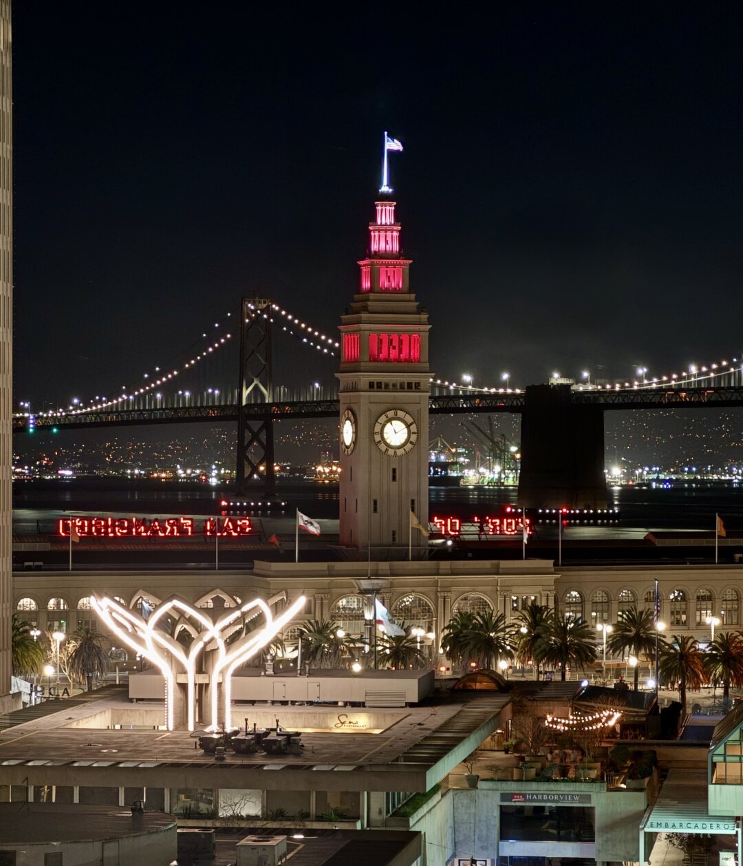 A view east at night from the San Francisco financial district showing the ferry building clock tower lit pink and red with the Bay bridge in the background